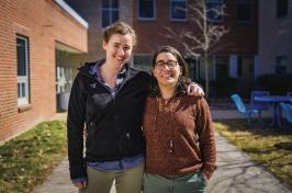 Two female graduate students stand outdoors with their arms around each other